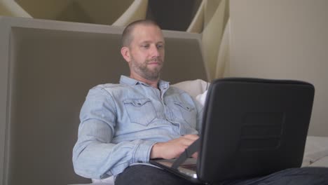 A-middle-aged-man-with-a-serious-and-focused-face-is-typing-and-working-on-his-laptop-from-the-bed-of-his-modern-hotel-room