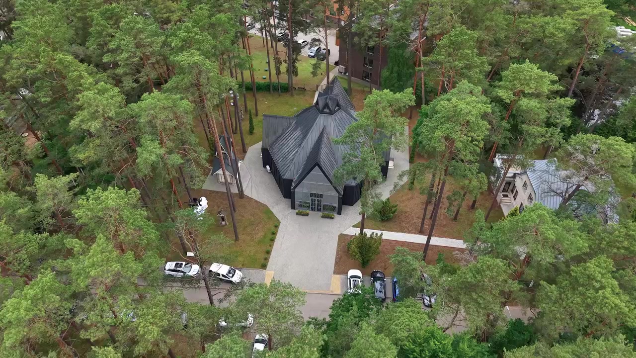 Ohromující dronové video ukazuje moderní kostel s černou střechou, uhnízděný v hustém borovém lese, obklopený venkovskými cestami, zaparkovanými auty a klidnou vesnicí za přirozeného denního světla.