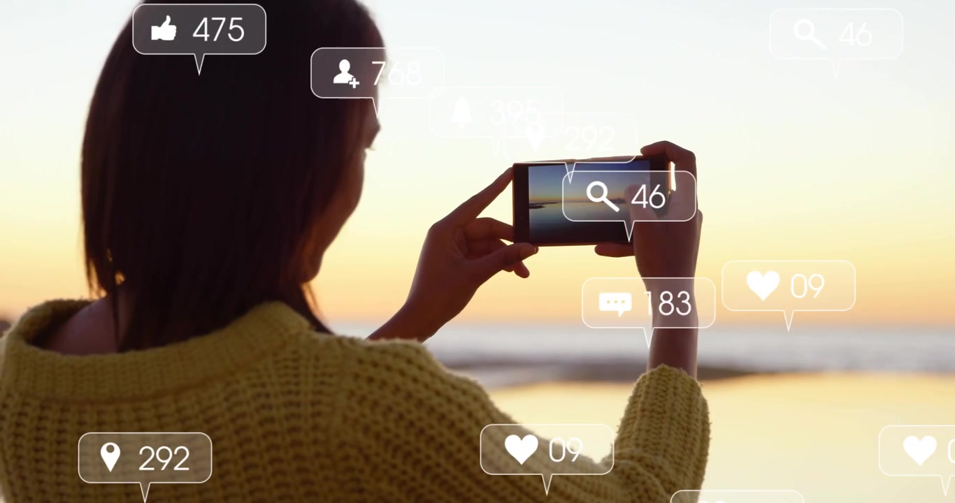 Woman raising phone at shore steadying and capturing sunset while social icons rising around phone