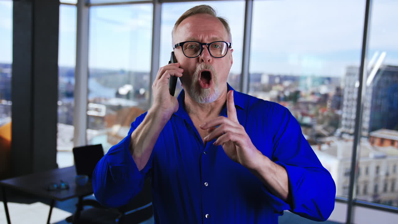 Relaxed mature man wearing blue shirt speaks on the phone in office. Man ends the conversation and kisses his phone.