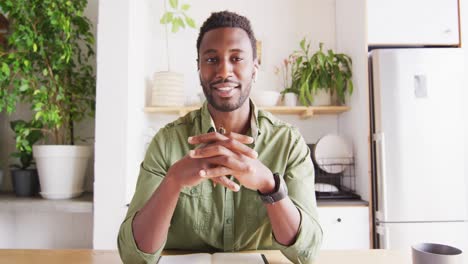 happy african american man sitting at table in kitchen, having video call