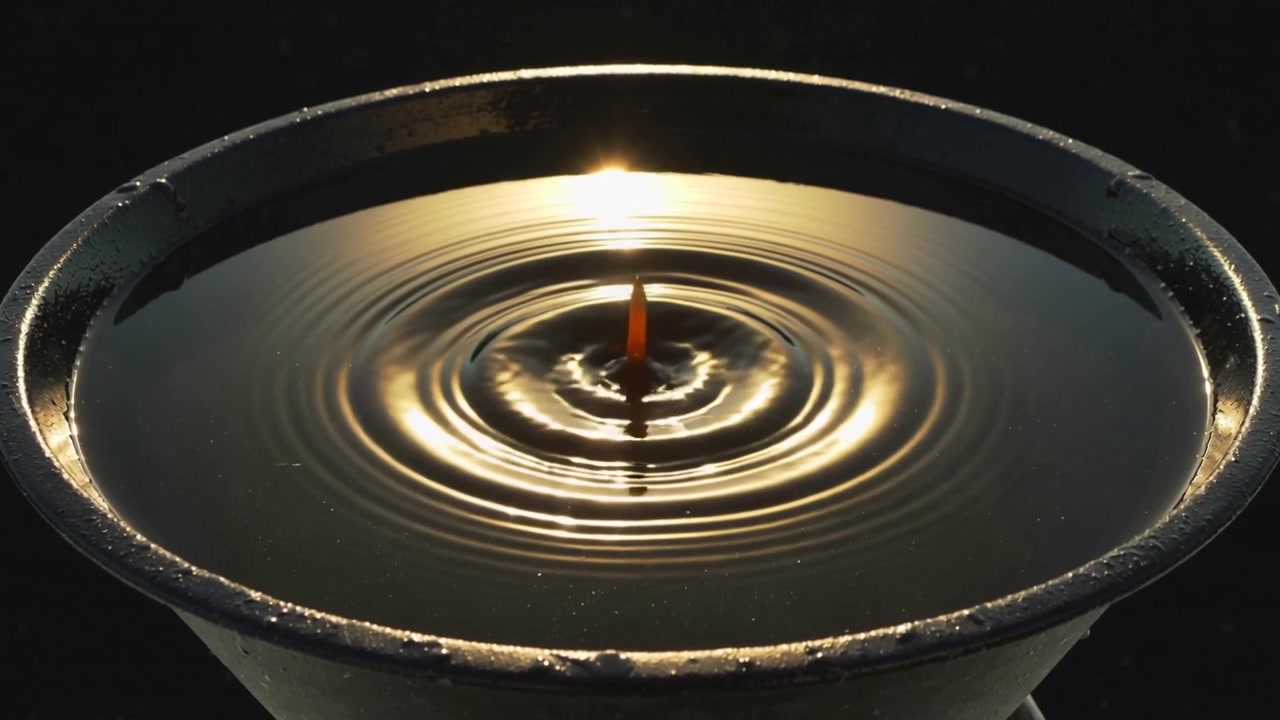 Dropping droplet hitting metal bowl filled with dark shiny liquid in studio creating golden ripples