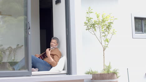 Hombre-Birracial-Senior-Enfocado-Sentado-Junto-A-Una-Ventana-Abierta-Tocando-La-Guitarra-En-Casa,-Espacio-Para-Copiar,-Cámara-Lenta