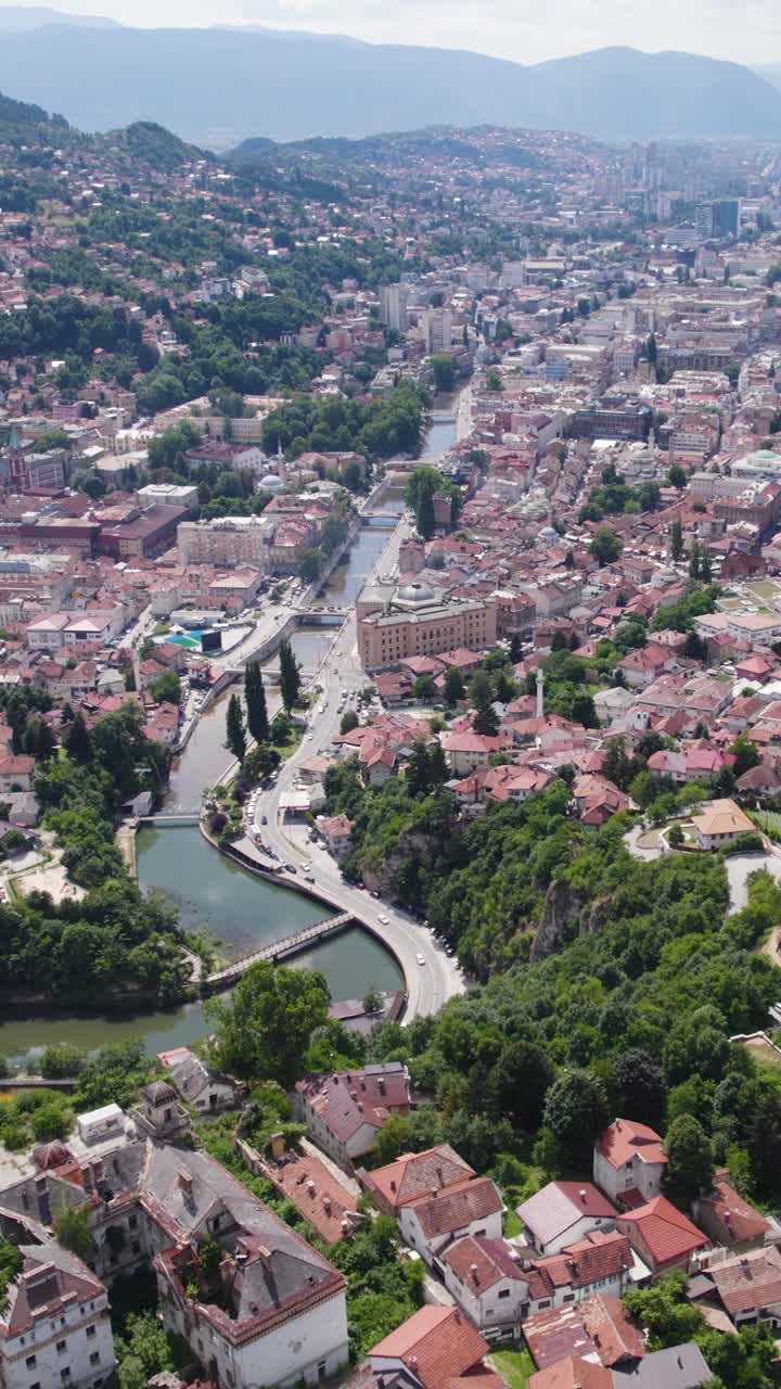Sarajevo showing dense rooftops, modern buildings, and green hill surrounds, vertical aerial pullback
