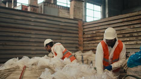 A-man-with-Black-skin-in-a-white-helmet-and-a-white-protective-uniform-in-an-orange-vest-walks-on-a-tramp-and-presses-cellophane-garbage-along-with-his-colleagues-a-man-with-a-beard-and-a-girl-in-a-blue-protective-suit