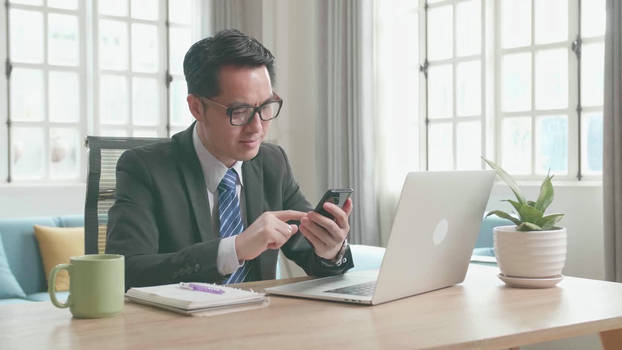 Asian Businessman With Glasses Wearing Business Suit Using Mobile Phone While Using Computer For Working At Home.
