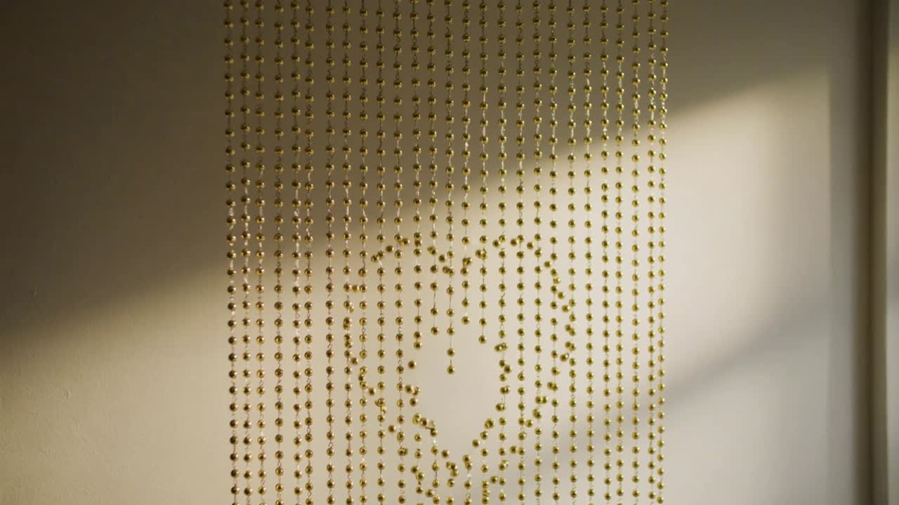 Shifting sunlight revealing heart gap in gold beaded curtain on pale wall at home, diagonal bands