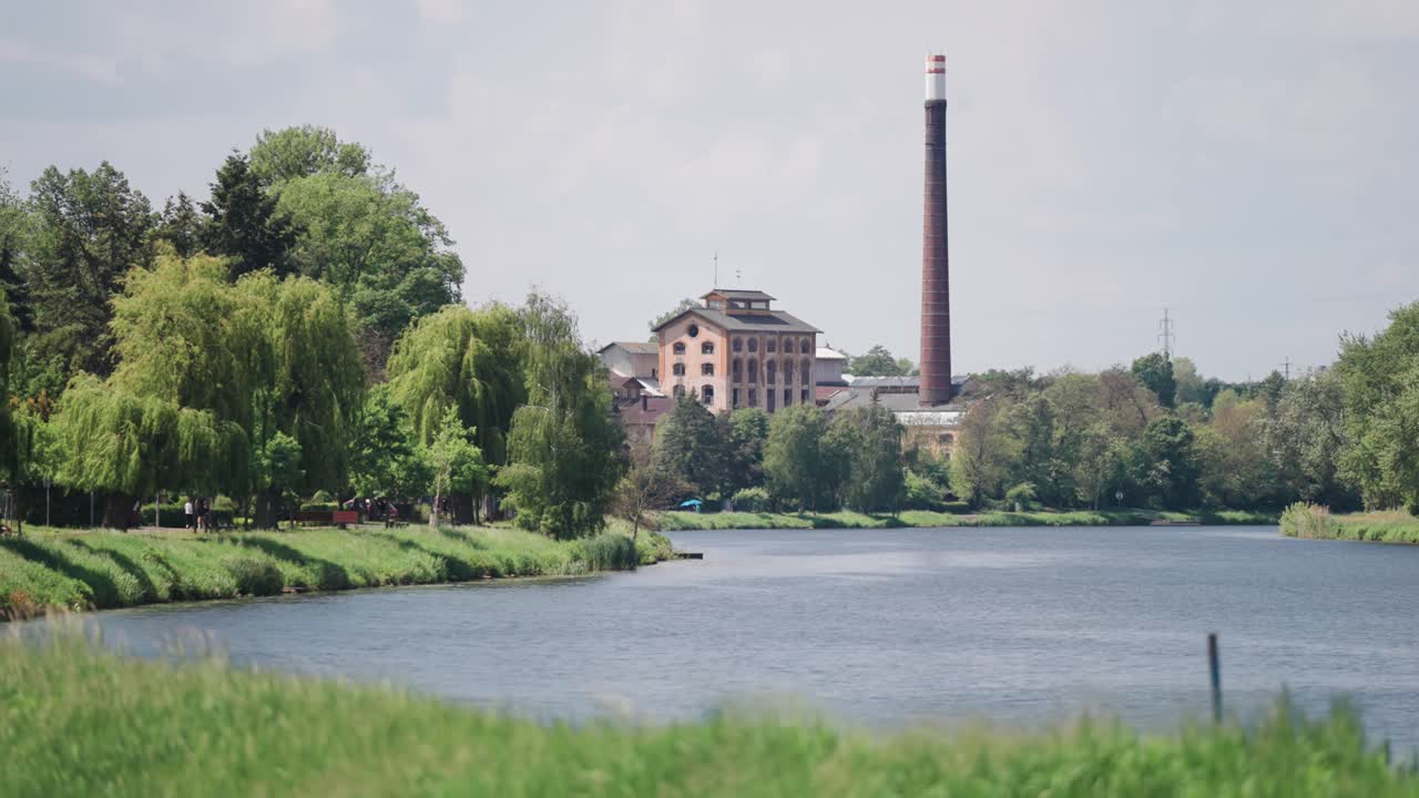 Industrial building with a tall chimney next to a river, surrounded by lush green trees.