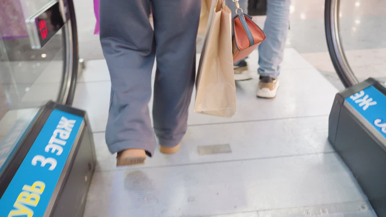 Close up leg view of shopper exiting escalator holding brown paper shopping bag, dressed in casual outfit, surrounded by mall environment, highlighting retail experience, and everyday lifestyle