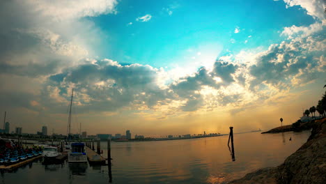 Long-Beach,-California-with-boats-docked-in-the-foreground,-the-city-skyline-in-the-background-and-a-colorful-sunset-reflecting-off-the-ocean---time-lapse