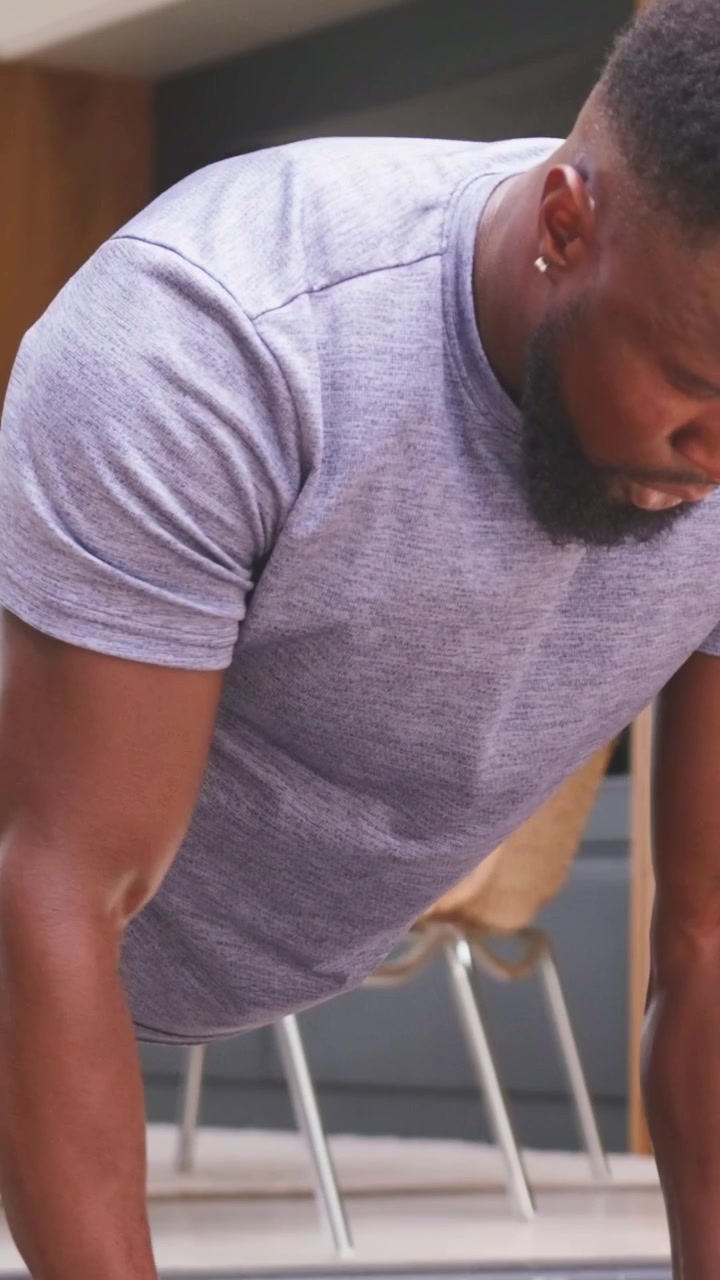 Vertical video: Starting plank, man in gray tee doing push-ups for home workout, gripping dumbbells