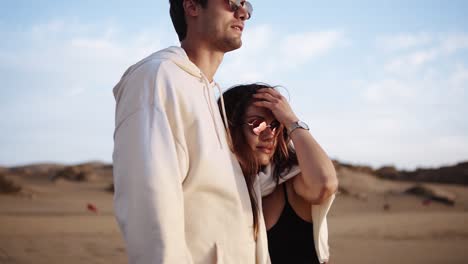 stylish, young couple standing on sandy beach shoreline watch sunset waves from the ocean. wearing sunglsses and casual clothes. front view