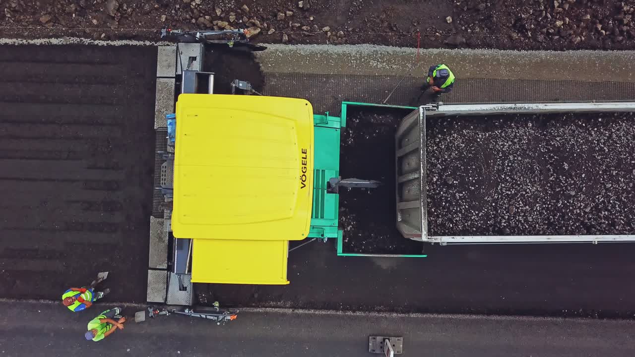 A big truck with bitumen and rubble pulls up to the paver on the background of a fresh asphalt strip and a few people around. Road repair. Aerial view.