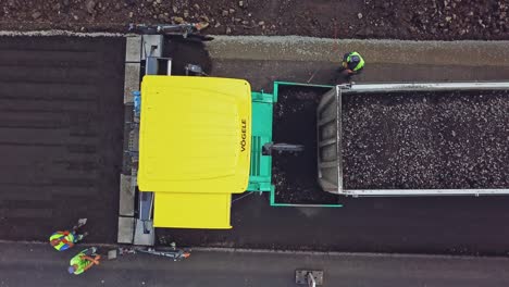 a big truck with bitumen and rubble pulls up to the paver on the background of a fresh asphalt strip and a few people around. road repair. aerial view.