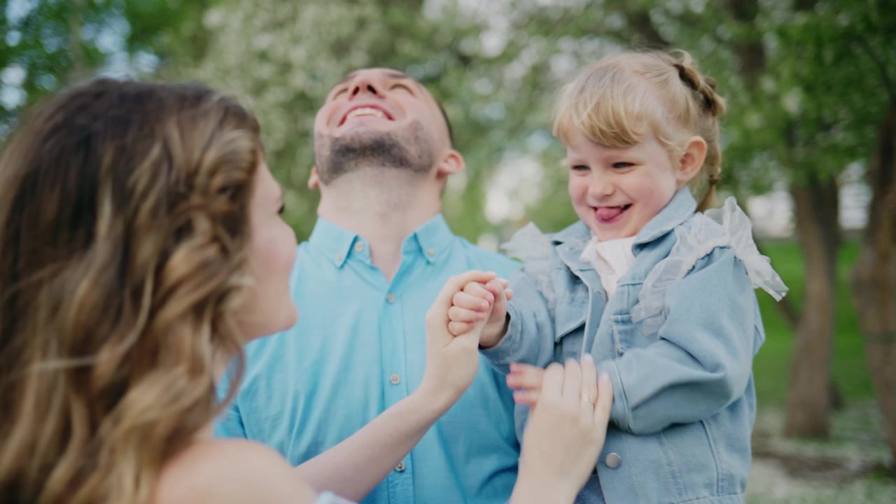 Una famiglia felice si gode una giornata di sole all'aperto. L'uomo sorride calorosamente mentre tiene in braccio il suo bambino che ride. La donna osserva con gioia affettuosa. Il bambino copre giocosamente il viso dell'uomo con le sue mani.
