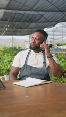 vertical video: african american man in apron talking on phone, smiling in greenhouse nursery