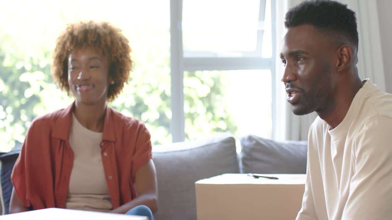 Diverse couple sitting on couch in living room, closed box sparking man speaking and gesturing
