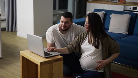 lovely pregnant woman and man surf the net sitting on the floor with laptop