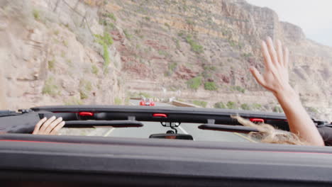 couple waving through roof of open top hire car on summer vacation