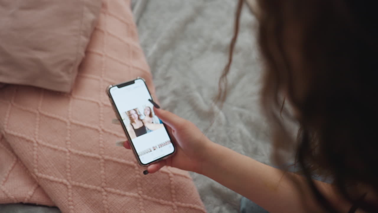 Overhead Shot Of Caucasian Woman Scrolling Memories On Smartphone While Seated On Pink Quilted Bed, Soft Daylight, Long Hair, Black Nails Tapping Portraits, Quiet Nostalgic Atmosphere And Casual