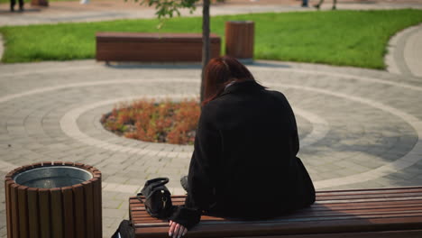 rear view of woman seated in public park with her hand resting on bench, handbag placed nearby, relaxed atmosphere with green surroundings and scenic park features in the background