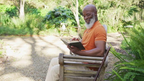 hombre mayor afroamericano sonriendo mientras lee un libro mientras está sentado en un banco en el jardín