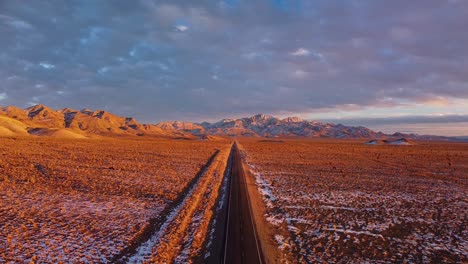 Antena-De-4k-De-Un-Dron-Que-Asciende-Por-Una-Carretera-Extraterrestre-Cerca-De-Rachel-Nevada-Que-Muestra-Una-Montaña-De-Tejones-En-El-Fondo-A-La-Hora-Dorada-Con-Nieve-En-El-Suelo-Y-Nubes-En-El-Cielo