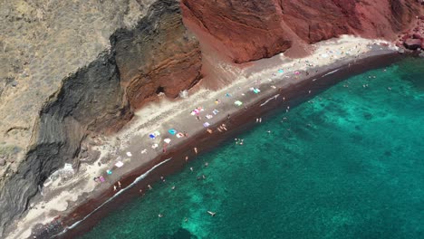 aerial top down rising and revealing red beach with turquoise water, tourists, mountains and red colored sand in santorini, greece