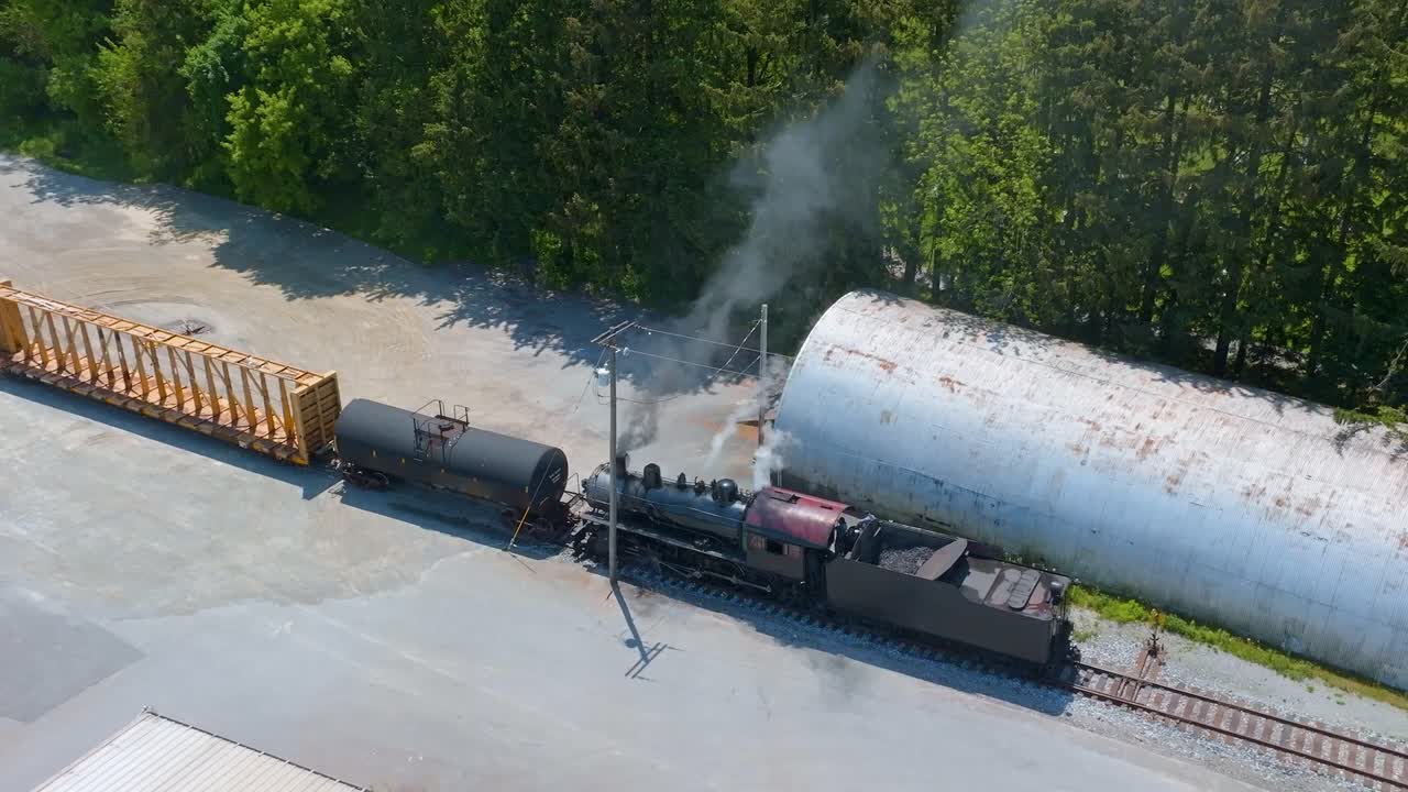 In a rural area, a vintage steam locomotive navigates railway tracks while pulling freight cars. Smoke billows from the engine as it moves, evoking nostalgia for classic rail travel.