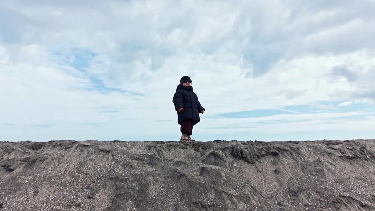 日中の屋外、ビーチまたは建設現場の近くで、砂利の山の上に立つ子供が曇り空を見上げている
