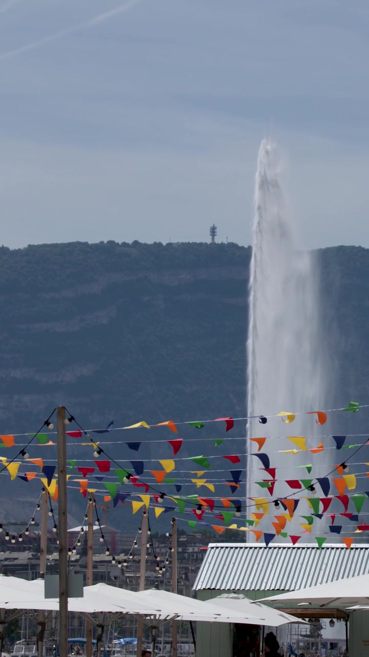 Inquadratura ampia statica di un enorme getto d'acqua che si innalza dietro bandiere di festa e montagne.