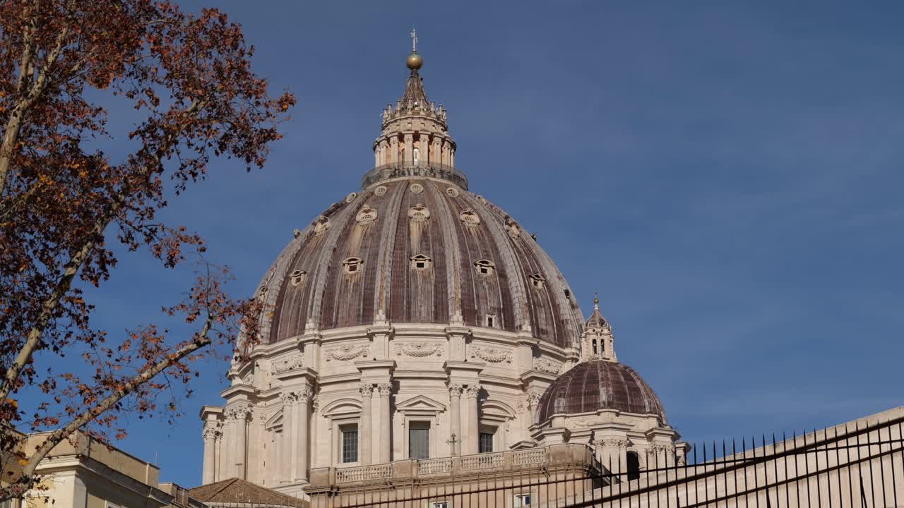 Detailed 4K shot of the dome of St. Peter’s Basilica in Vatican City, Rome, framed with autumn tree branches against a bright blue sky, highlighting historic architecture