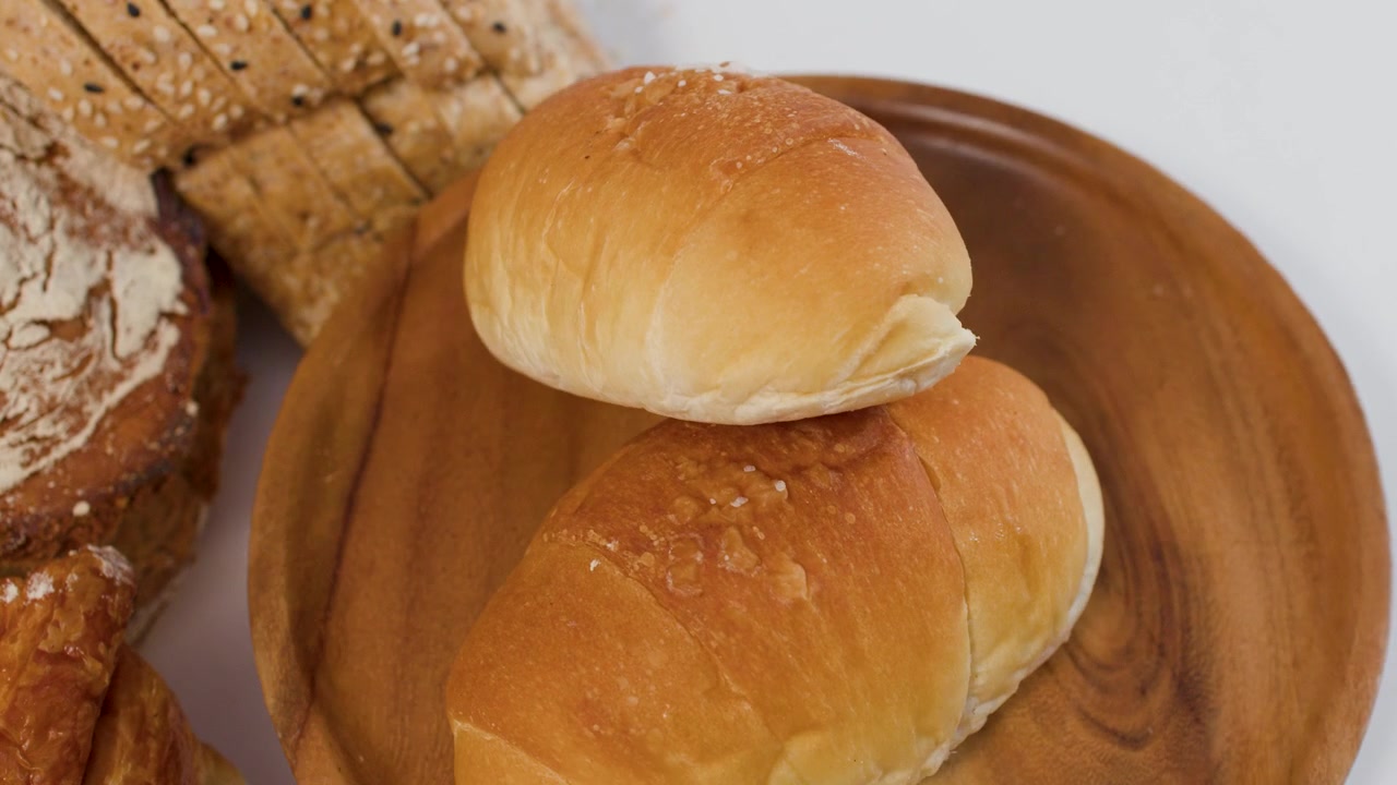 Rotating wooden plate holding golden dinner rolls beside sliced sourdough and baguette on white background.