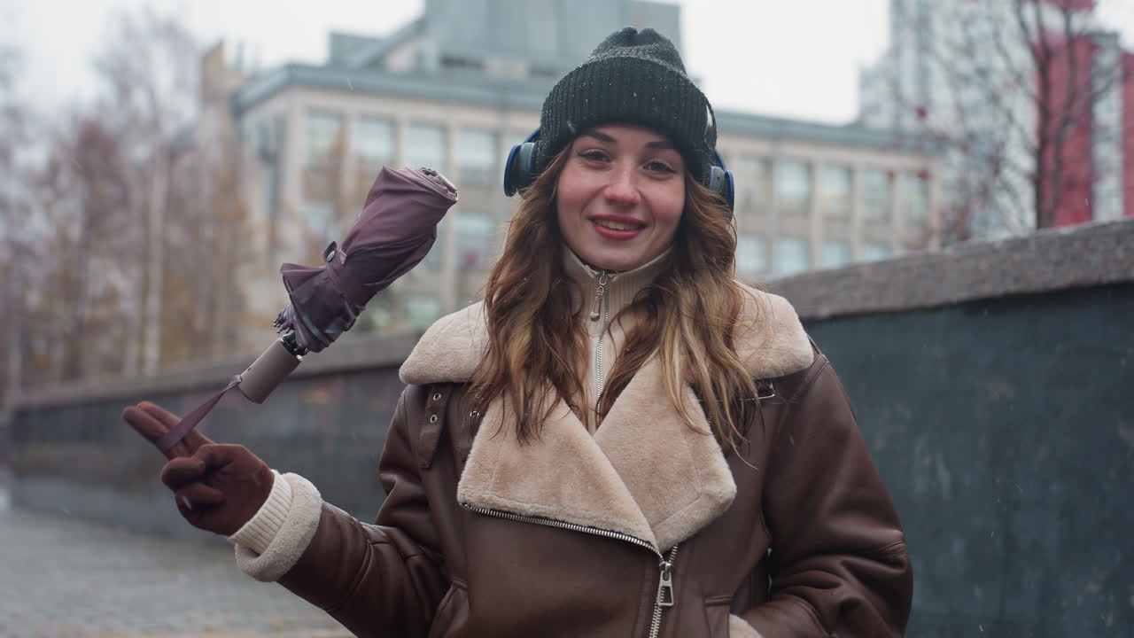 Portrait shot of woman in black knit cap, brown shearling jacket, standing outdoors on concrete path covered with autumn leaves, smiling and swinging umbrella playfully during chilly weather