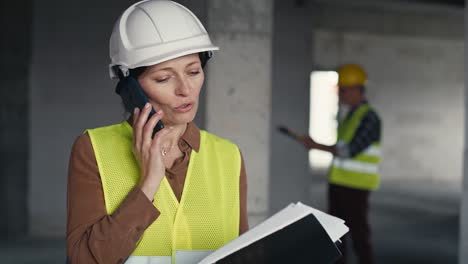 Waist-up-video-of-female-caucasian-engineer-on-the-construction-site-talking-on-the-phone-and-checking-some-documents.