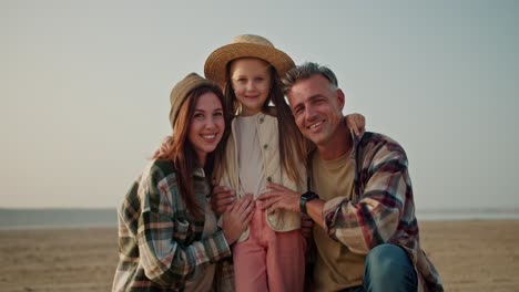 Portrait-of-a-happy-family,-a-little-blonde-girl-in-a-Straw-hat-stands-in-the-middle-and-is-hugged-from-different-sides.-Her-father-is-a-brunette-man-with-gray-hair-in-a-checkered-shirt-and-her-mother-is-a-girl-and-a-brunette-in-a-Green-checkered-shirt-on-a-deserted-shore-during-a-summer-hike-outsid