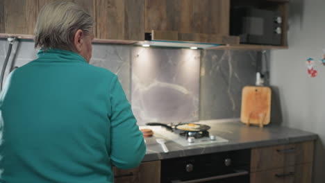 older woman standing at stove in modern kitchen, cooking eggs in frying pan, with kitchen countertop and utensils visible, focused on meal preparation process in home cooking setting