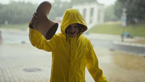 Cheerful-teenage-girl-in-a-yellow-jacket-with-a-hood-pours-water-from-a-rubber-boot-while-raining-and-walking-in-the-park