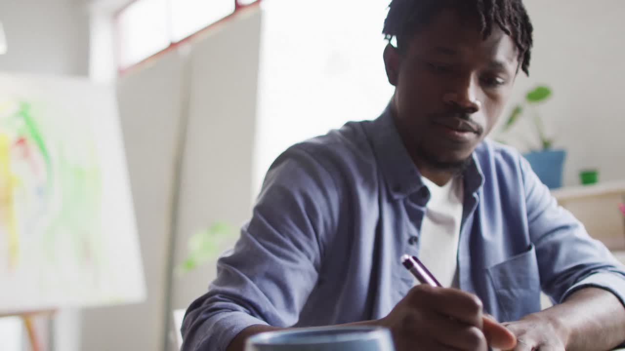 Artista uomo afroamericano che prende appunti mentre è seduto alla sua scrivania in uno studio d'arte.