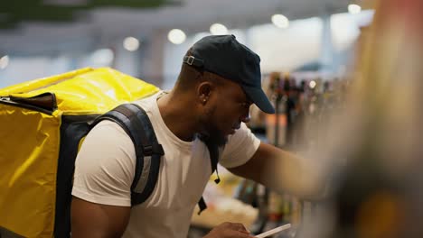 A-Black-skinned-food-delivery-man-with-a-large-yellow-bag-selects-the-necessary-goods-from-among-the-products-in-a-grocery-store-window.-A-delivery-man-checks-the-necessary-items-and-buys-them-to-deliver-with-a-large-yellow-bag