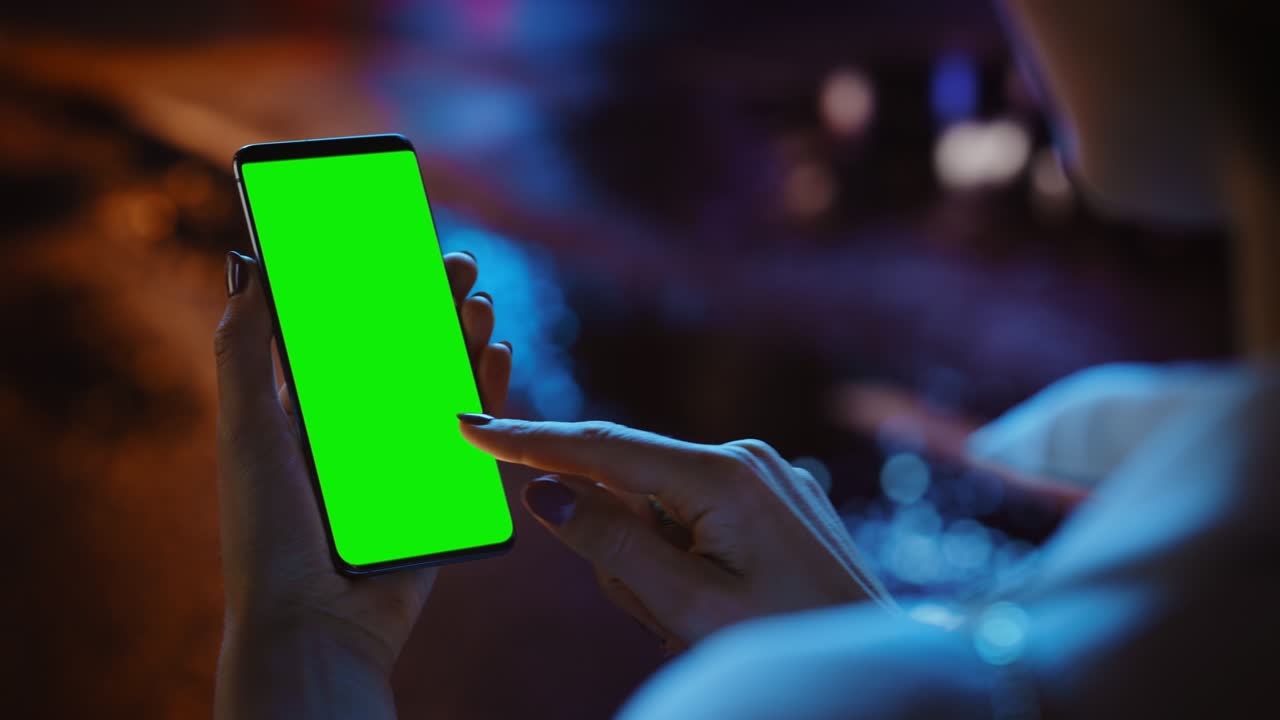 Woman using Chroma Key Smartphone while Standing, Gesturing, Swiping, Scrolling. Night City Street Full of Neon Light. Female Using Green Screen Mobile Phone. Over the Shoulder Close-up Shot