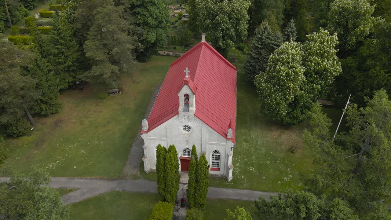 Aerial ascending view of Rannamõisa limestone red roofed church surrounded by vibrant green foliage and rural churchyard. Drone perspective of Estonia countryside village chapel and lush canopy around