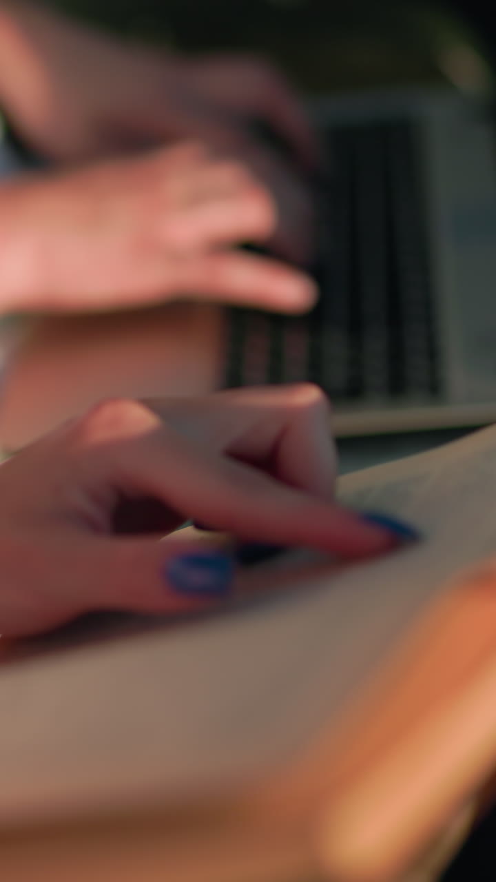primer plano de una mujer de color rosa sosteniendo un libro, interactuando con su amiga escribiendo en una computadora portátil cercana, centrarse en las manos y el libro, fondo borroso, luz natural cálida