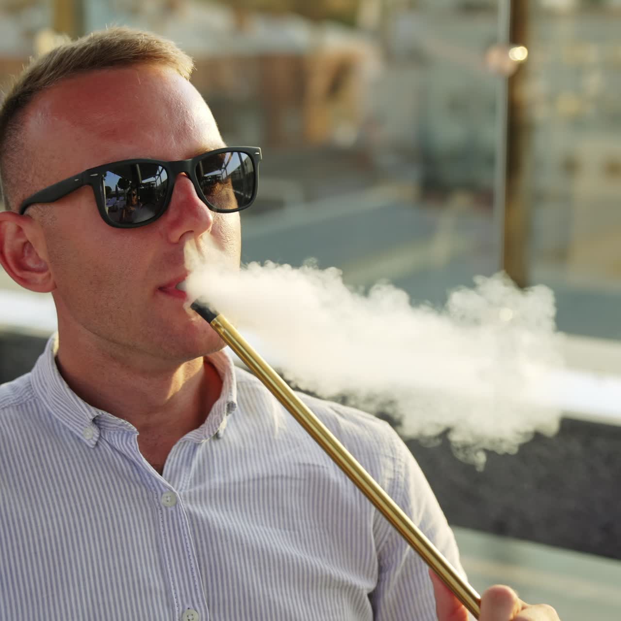 Mid aged man wearing sunglasses and shirt sits outdoors. Male resting and smoking hookah producing a cloud of thick white smoke