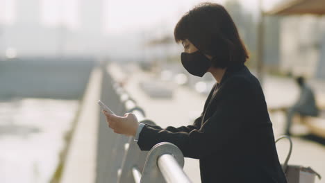 asian businesswoman in mask using phone on urban embankment