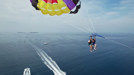 couple on summer vacation parasailing from boat over blue sea, drone dolly in towards people, then follow locked in