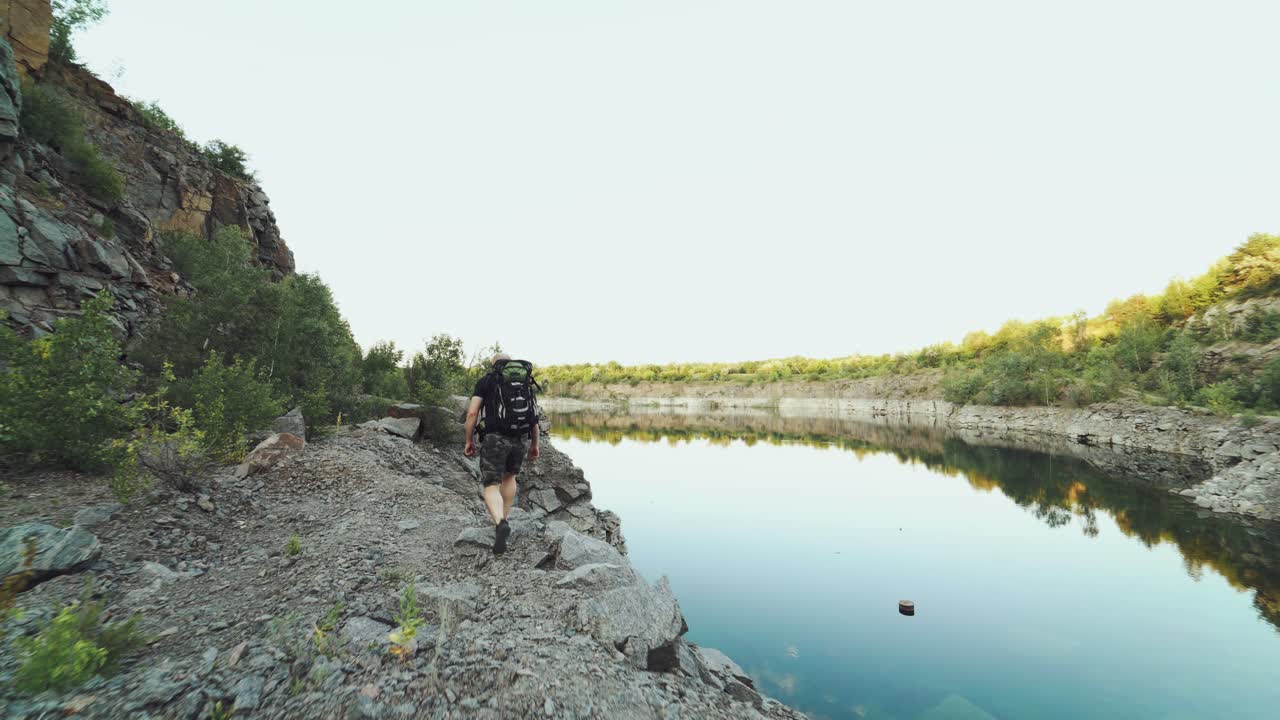 tourist with a backpack on his back is going along the bank of the lake on the background of high rocks and wild nature on a summer day