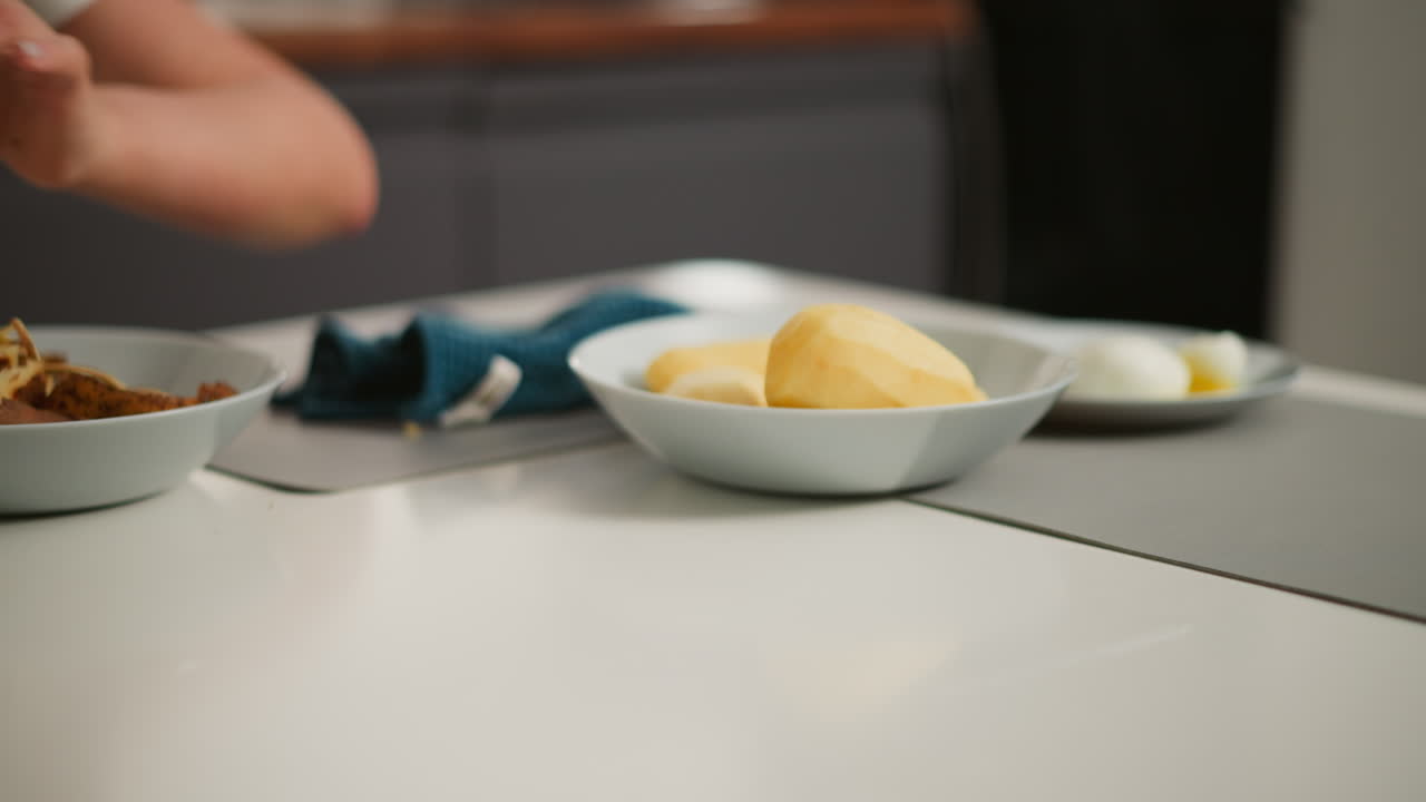 Partial view of person placing peeled potato into bowl using hand holding yellow peeler, with nearby plate containing white boiled eggs on light-colored kitchen counter during meal prep