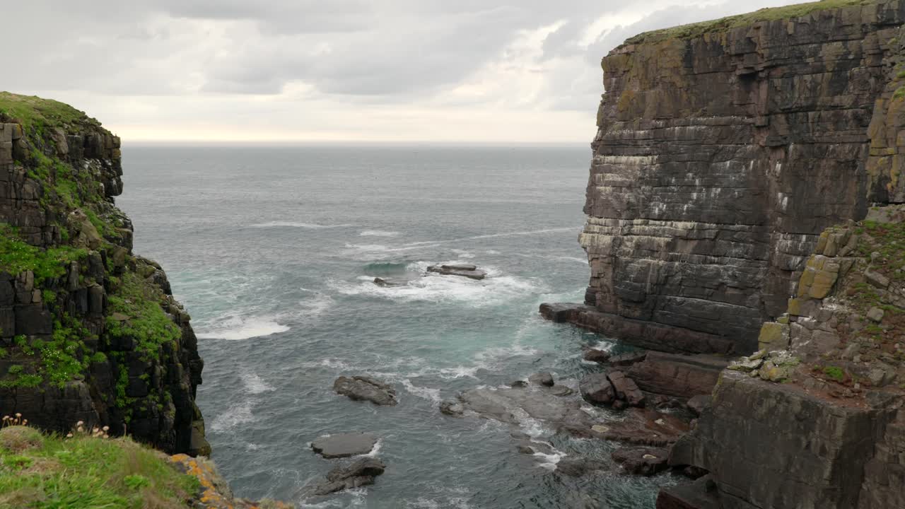 Looking out from a bay, waves crash against a tall and imposing sea cliff in the ocean while seabirds fly around the cliffs of a seabird colony of guillemots and puffins on Handa Island, Scotland