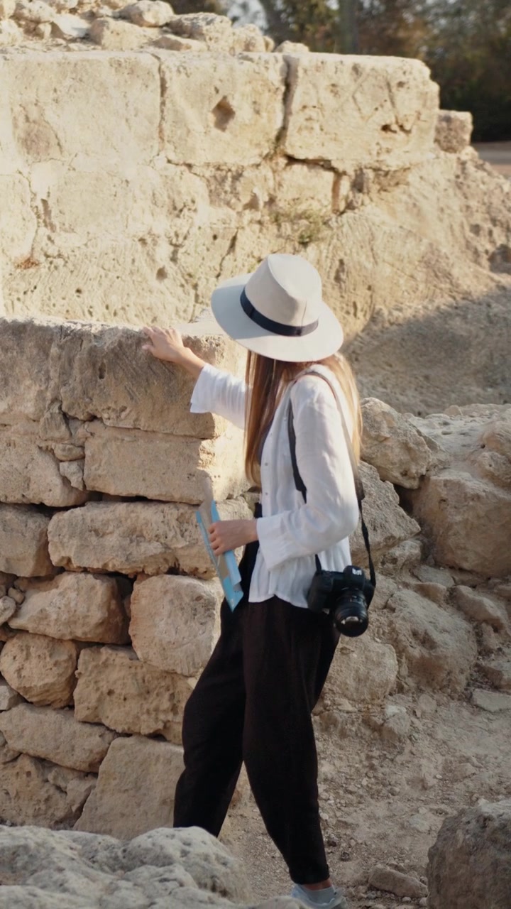 Una donna con una macchina fotografica esplora antiche rovine indossando un cappello e una camicia bianca, video verticale